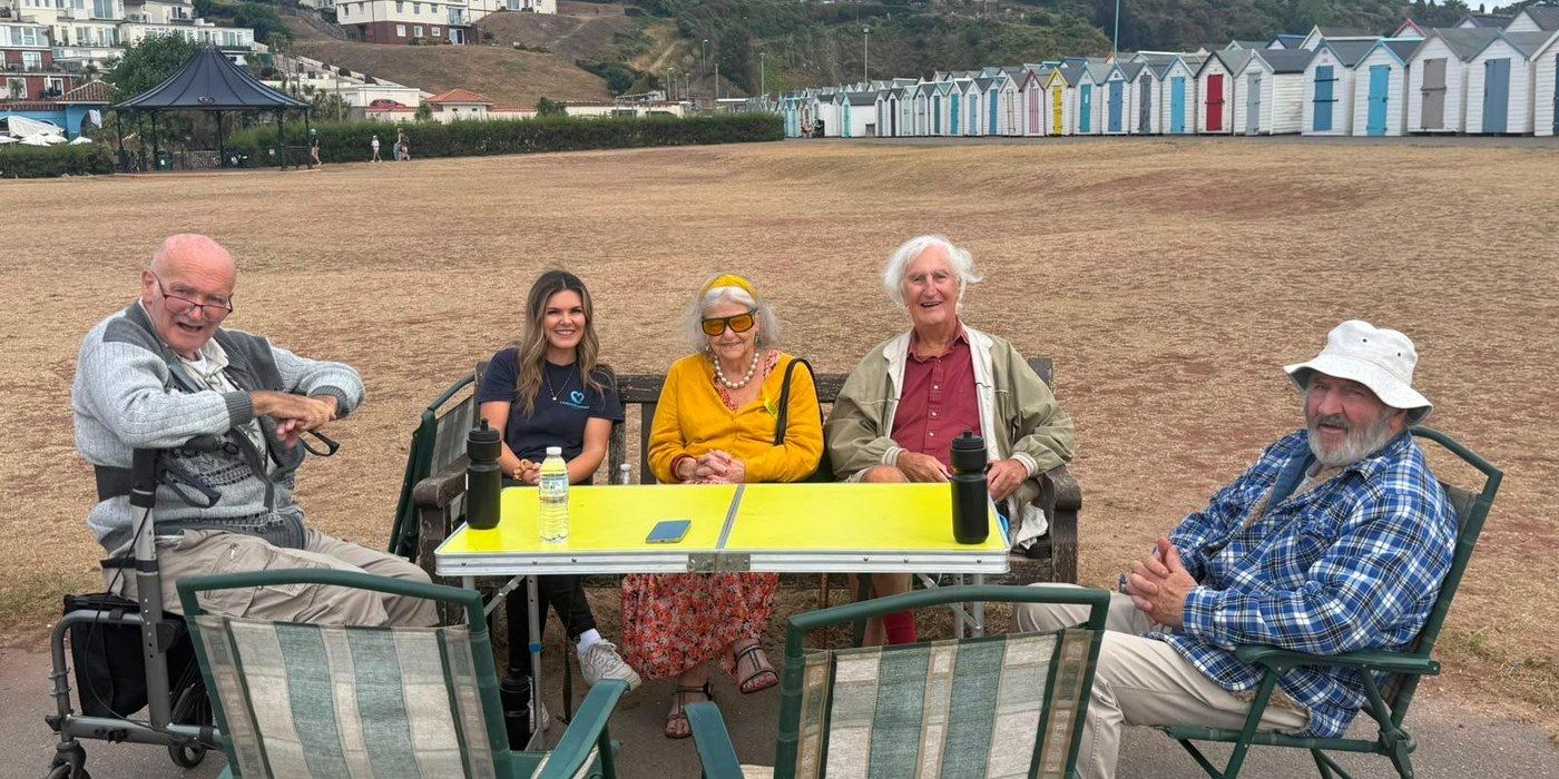 Five guests and a member of staff sitting round a folding table on the green at Broadsands, with the beach huts behind