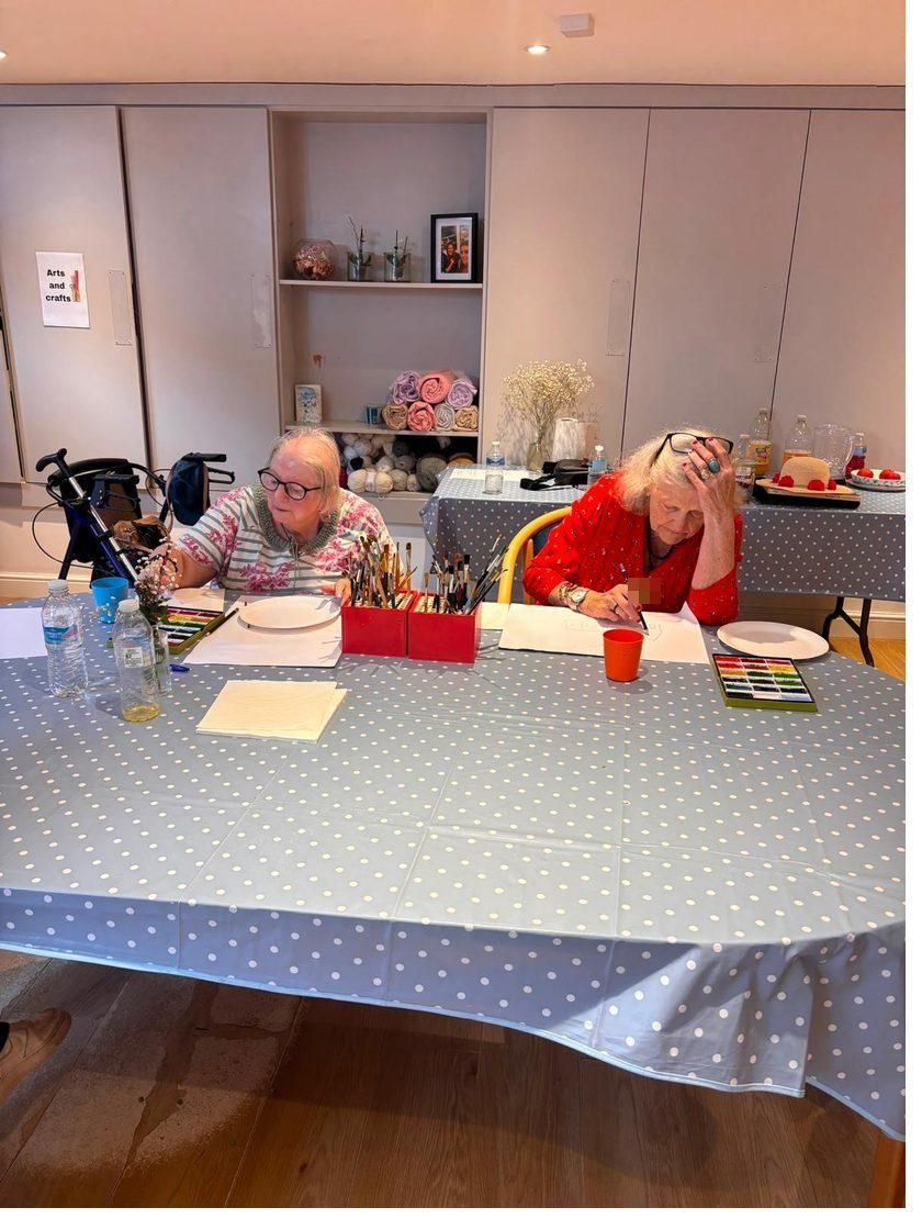Two ladies painting with watercolours at the craft table, brushes in a pot between them